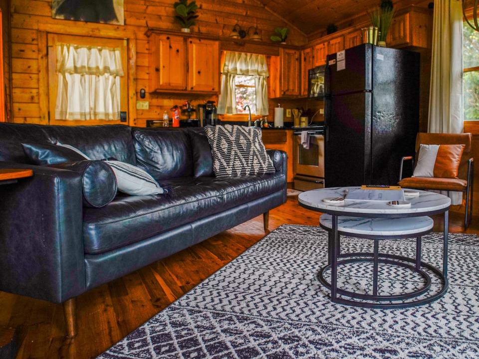 A leather black couch sits on the left side behind a neutral, geometric rug and two circular stacked white coffee tables, and wooden floors. Kitchen is seen in the background.