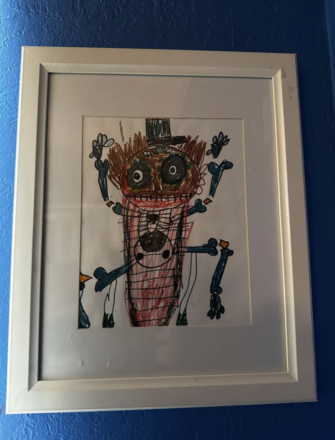 Child's drawing of a whimsical figure with a top hat, framed and hung on a blue wall