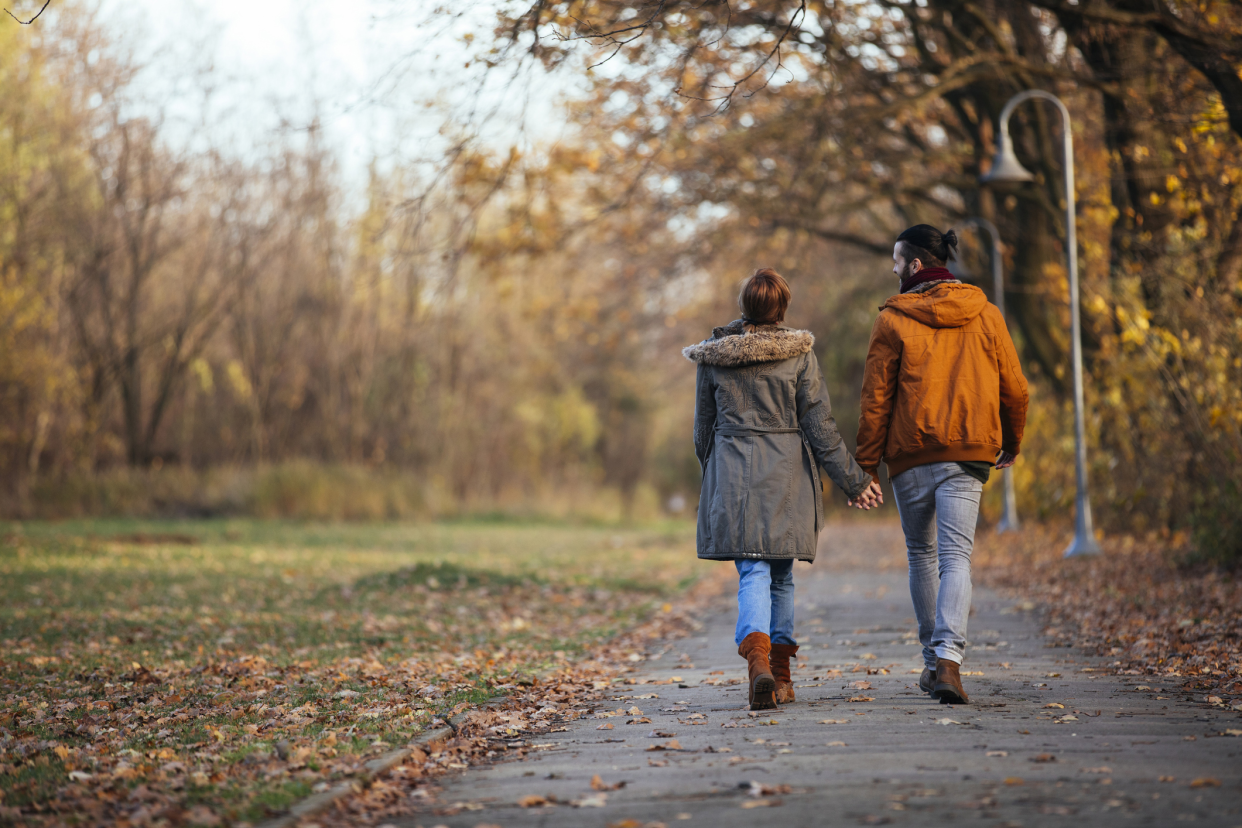 Young couple walking on a walkway in a park during late autumn with lots of orange and yellow leaves on the grass, both are wearing coats with a blurred background of trees