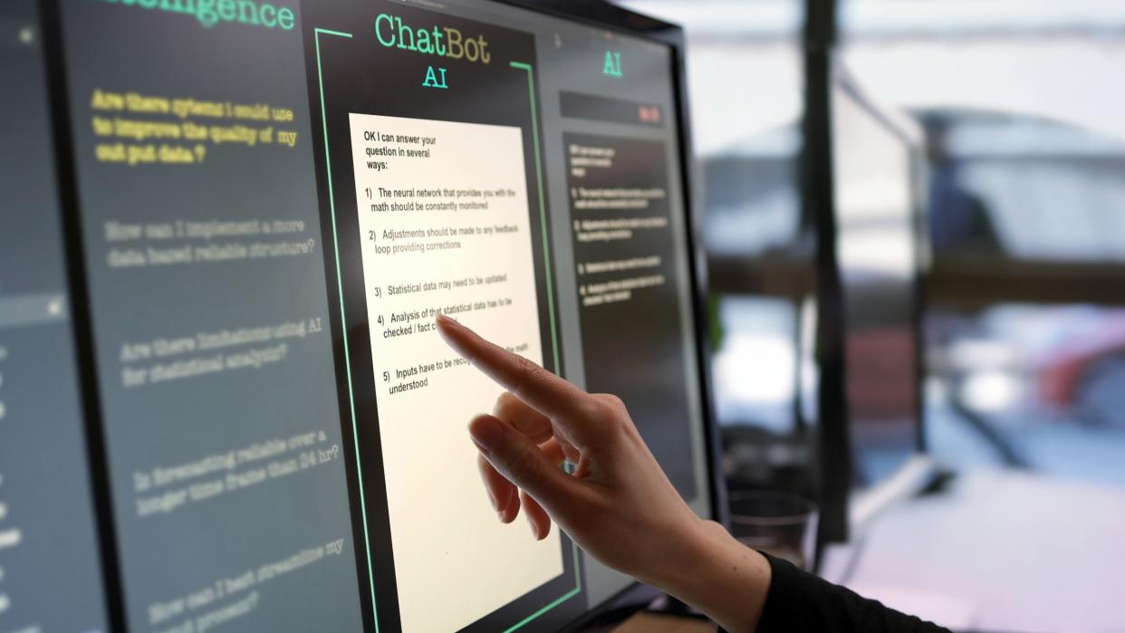 Close-up stock photograph showing a touchscreen monitor being used in an open plan office.A woman’s hand is asking an AI chatbot pre-typed questions & the Artificial Intelligence website is answering.