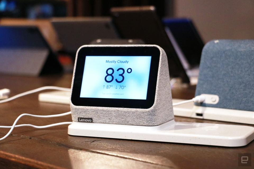 <p>Off angle view of a gray Lenovo Smart Clock 2 on a wireless charging dock with its screen facing the camera. In the background sits a blue Smart Clock 2 and a row of tablets.</p>
