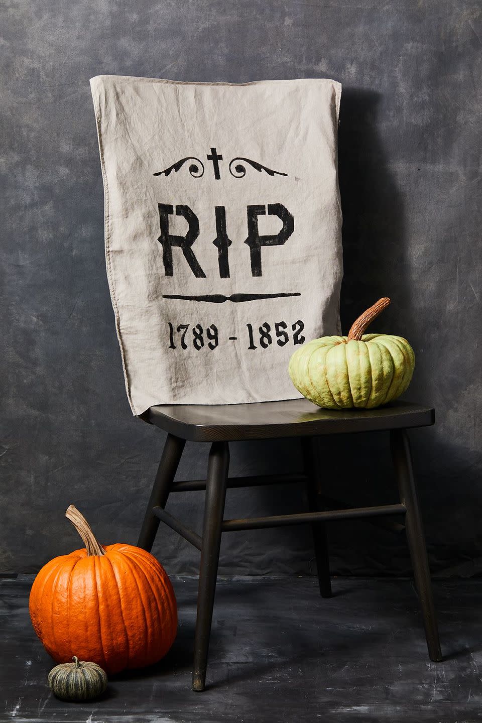 <p>Your guests will be afraid to sit down at their seat once they see these creepy chair covers. To make, <a href="https://www.goodhousekeeping.com/holidays/halloween-ideas/a33564450/gh-halloween-artwork-calligraphy-templates/" rel="nofollow noopener" target="_blank" data-ylk="slk:print out the tombstone templates;elm:context_link;itc:0;sec:content-canvas" class="link ">print out the tombstone templates</a> and cut out designs with scissors or a craft knife so you can use them as stencils. Place your template centered on your pillow case in the position that feels right and tape or pin the corners down to keep in place. Dip a foam craft brush in a small amount of black fabric paint and dab it gently onto the fabric where you have placed the stencils (less is more!). Let dry completely. Optional: Add some extra dabs of gray fabric paint on top of the dried black designs to give it a distressed look. Slip pillowcases over the back of your dining chairs. </p>