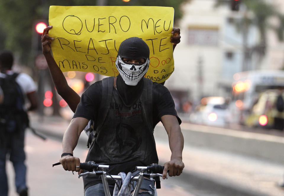 A masked protester rides a bike as his passenger holds a sign that reads in Portuguese; "I want a readjustment," in reference to the increased bus fare, protesting not only against a 10-cent hike in bus fares but demanding they lower the price from the previous fare, in Rio de Janeiro, Brazil, Thursday, Feb. 13, 2014. Some roads in the center of Rio, which will serve as the 2016 Olympic city, were blocked Thursday night as demonstrators carried banners calling for a reversal of the fare hike, along with more investments in education and health care. (AP Photo/Silvia Izquierdo)