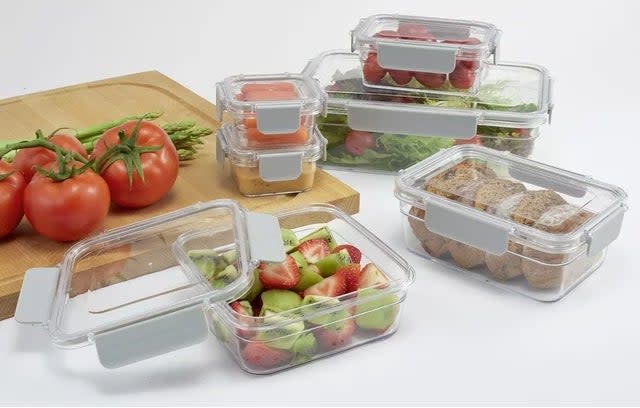 Assorted clear food storage containers with fresh produce and sandwiches inside, displayed on a table with tomatoes and a cutting board nearby