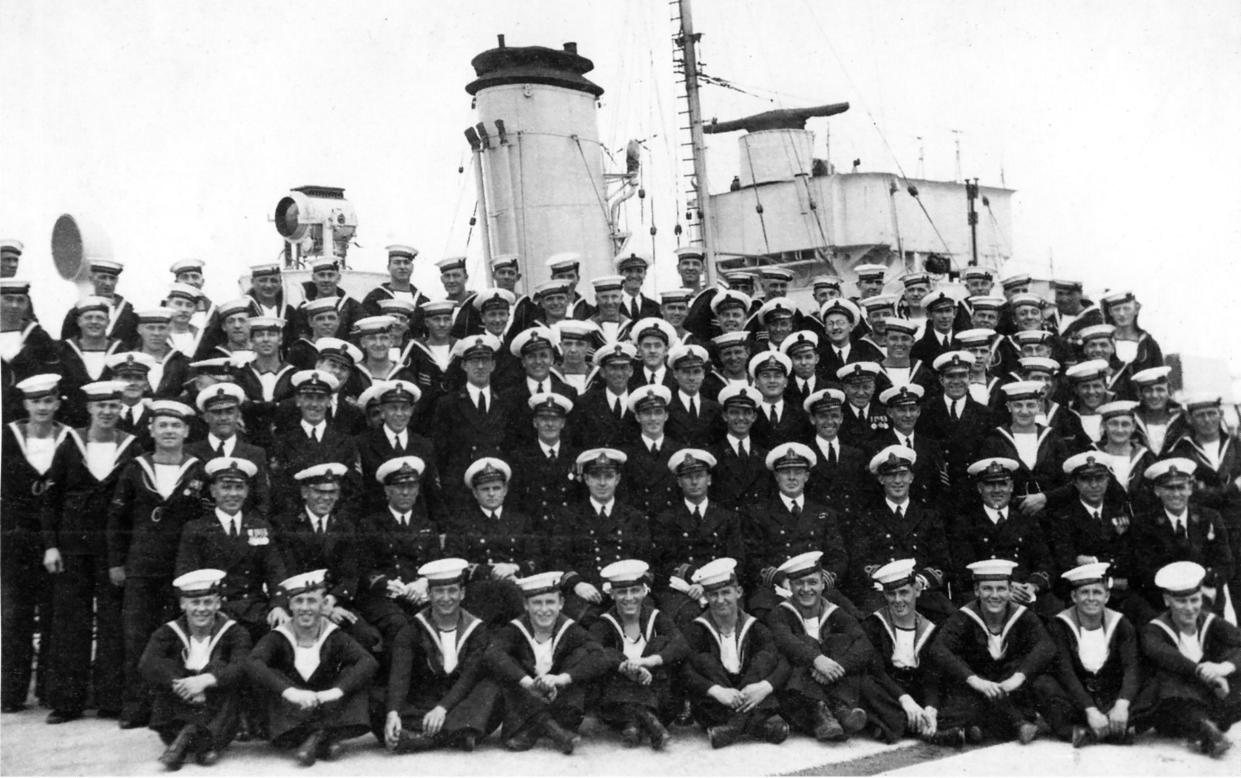 The black and white photograph shows many sailors sitting in a group portrait with the funnel of the ship visible behind