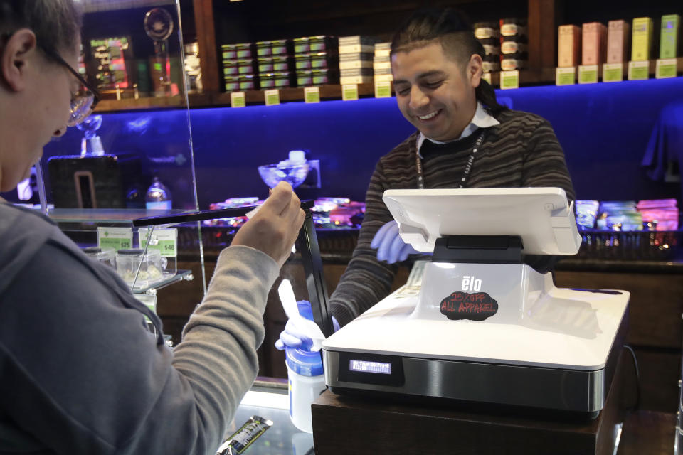 Max de Leon, right, a budtender at The Green Cross cannabis dispensary, smiles while handing a cleaning wipe to a customer in San Francisco, Wednesday, March 18, 2020. As about 7 million people in the San Francisco Bay Area are under shelter-in-place orders, only allowed to leave their homes for crucial needs in an attempt to slow virus spread, marijuana stores remain open and are being considered "essential services." (AP Photo/Jeff Chiu)