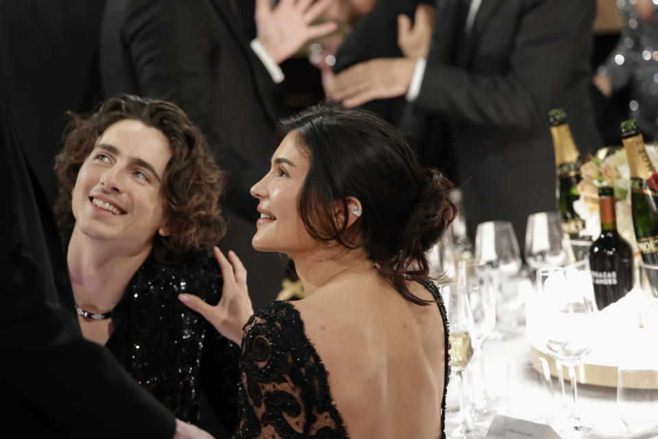 Timothée Chalamet and Audra Mari seated at a formal event, smiling and looking at someone out of the frame, surrounded by others in evening attire