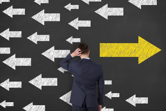 A man scratching the back of his head while facing a wall on which are drawn arrows going in different directions.