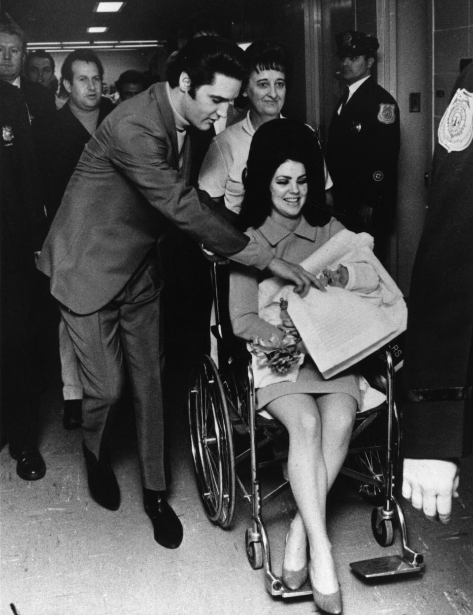 Elvis leaning over Priscilla, who's in a wheelchair and carrying baby Lisa-Marie