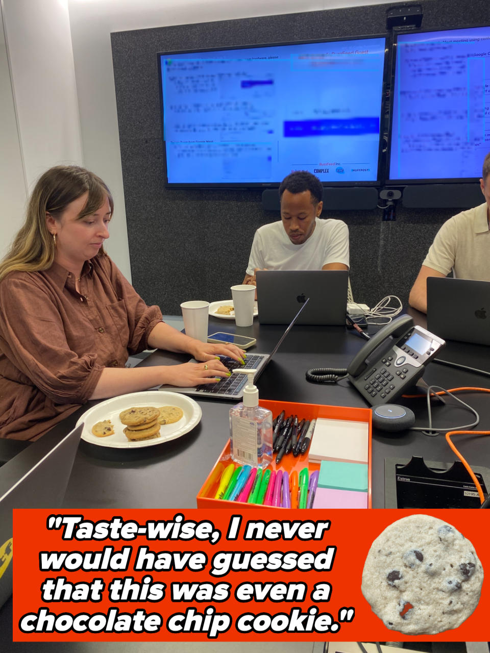 Three people working on laptops with snacks and office supplies on a conference table. Screens in the background display a presentation