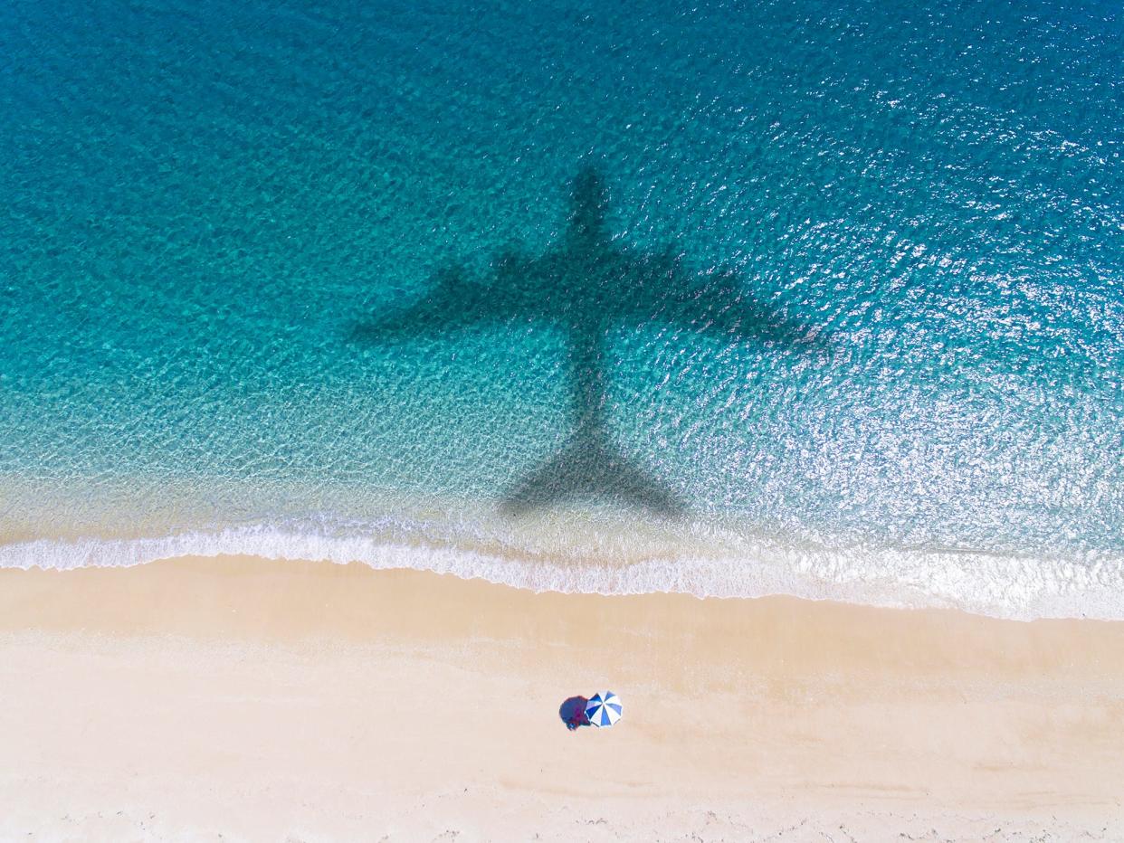 The silhouette of an airplane above a beach, with a single beach umbrella on the sand.