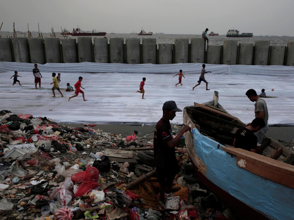 Fishermen repair their wooden boat, as children play football near the construction site of a concrete sea wall, in the Cilincing district of Jakarta