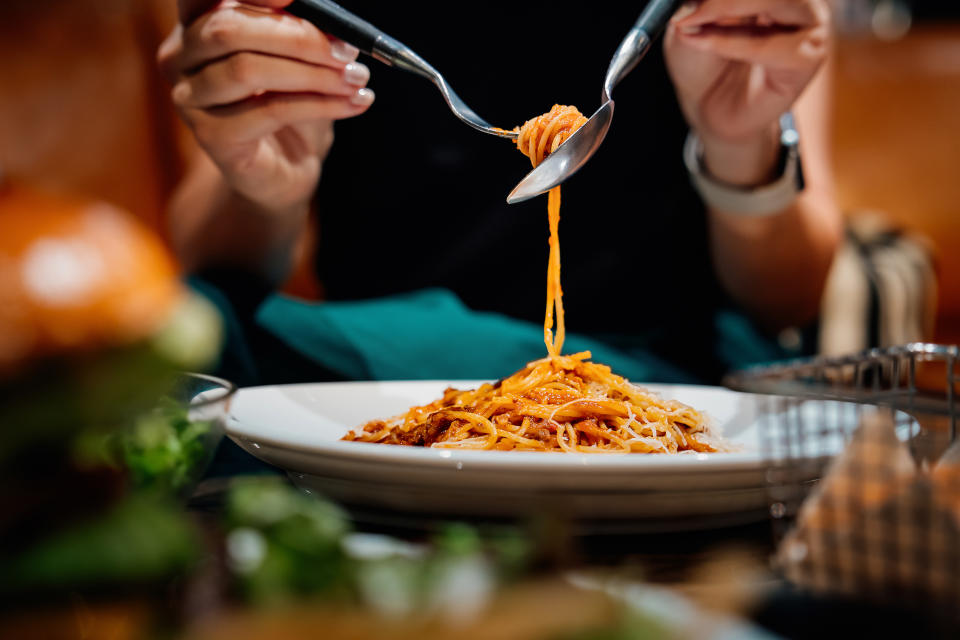 Close-up of a person eating spaghetti with a fork and spoon in a restaurant.  The background includes a blurred image of a hamburger