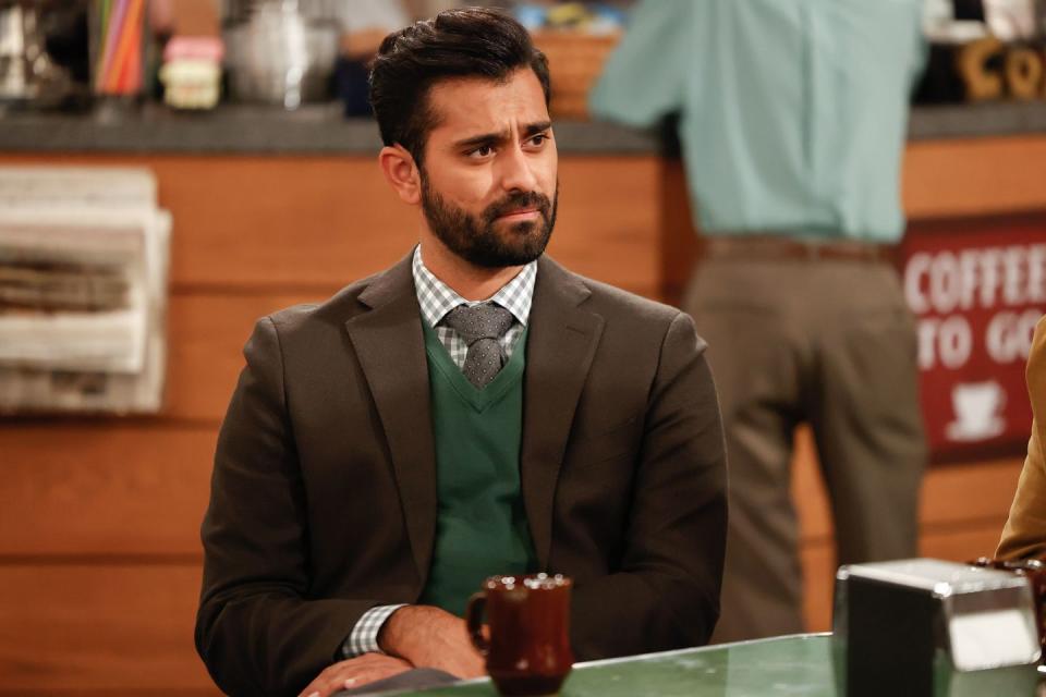 kapil talwalkar, an actor with dark hair and a dark beard, wearing a checkered shirt, gray tie, green sweater, and brown blazer in character as neil in the nbc show night court