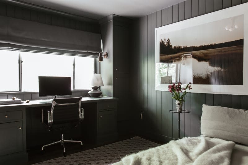 dark green tongue and groove wall paneling, pond landscape with train in background, small metal side table, small vase with flowers, low beige day bed, black and white checkered tile floor, desk build in under window