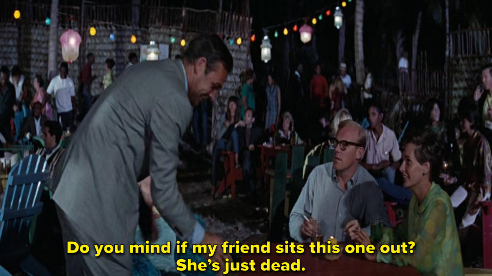 James Bond helps sit the corpse of a woman at a table while shocked guests look on