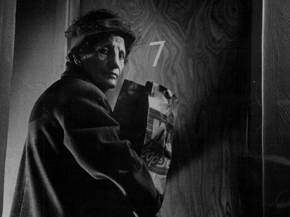 An older woman holding a paper bag looks over her shoulder as she enters her apartment.