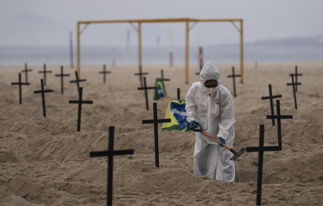 A person wearing white coveralls with a hood and face mask digs a hole on a beach surrounded by a few dozen black crosses upright in the sand as though marking graves.