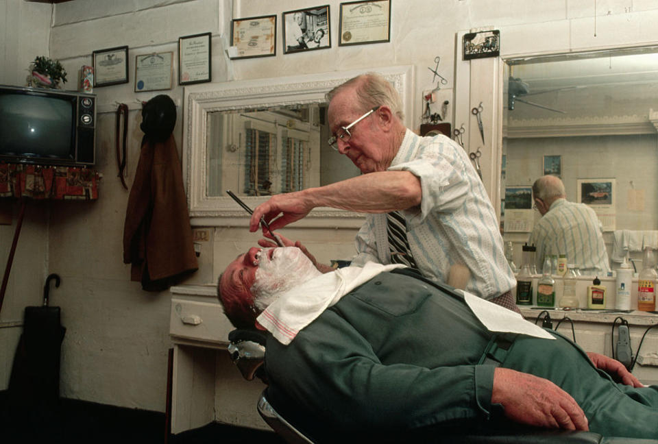 Barber shaves a man in a vintage barbershop, surrounded by framed certificates and a mirror. The setting reflects a classic, nostalgic atmosphere