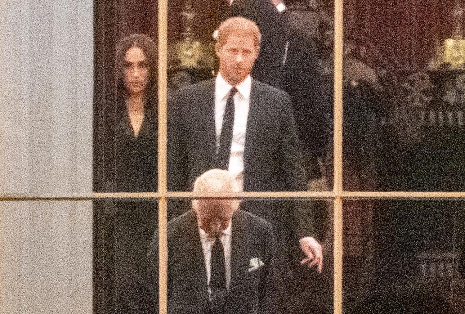 A close-up photo shows Meghan Markle, Prince Harry, and King Charles through a window of Buckingham Palace.