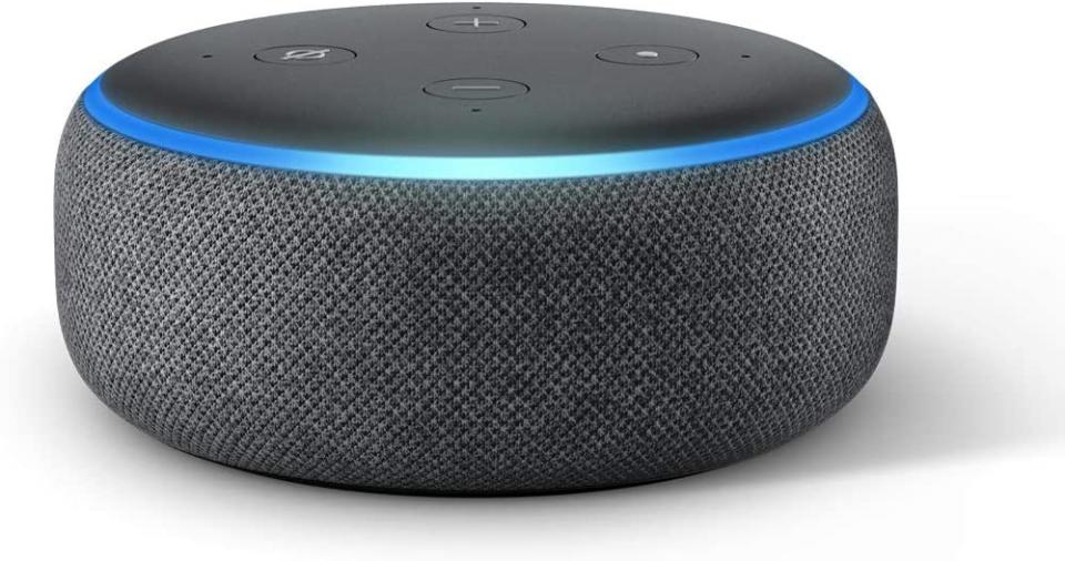 A round cushion shaped Echo dot device with electronic controls on the top, in grey shades with blue trim.