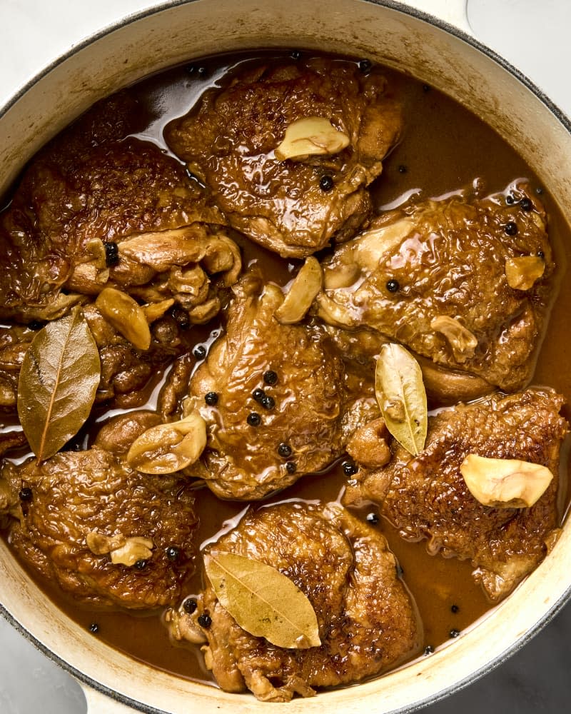 A dutch oven full of chicken thighs cooked in Filipino adobo with peppercorns and bay leaves visible.