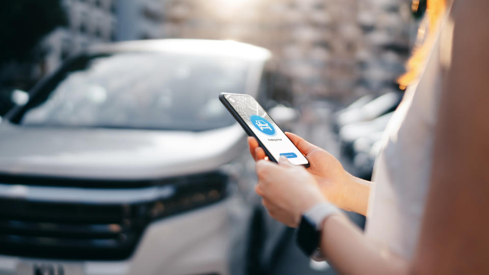 Cropped shot of young woman using mobile app device on smartphone to arrange a car pick up service in downtown city street, with busy city traffic and city scene as background