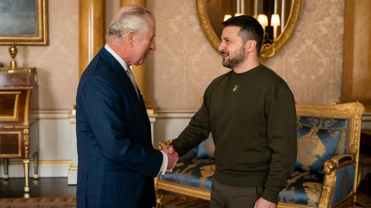 Two white men shake hands. THe one on the left is wearing a deep blue suit, while the one on the right is wearing a long-sleeve olive shirt. 