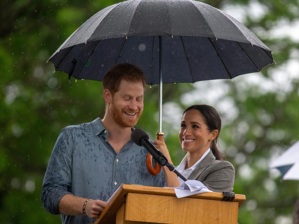 Markle holding an umbrella over herself and Harry as he speaks at a podium and it rains around them.