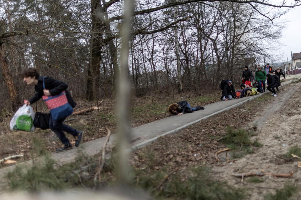 Ukrainians dressed for cold weather, some standing and some on the ground, look for cover on a sidewalk partially covered by bare trees.