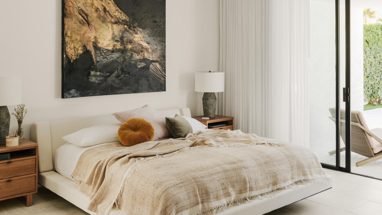  An off-white bedroom with neutral upholstered bed, beige throw, round terracotta pillow, sheer curtains and artwork above bed 