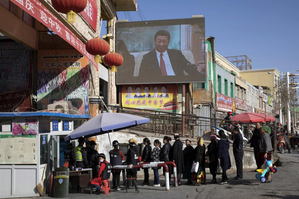 FILE - Residents walk through a security checkpoint into the Hotan Bazaar where a screen shows Chinese President Xi Jinping in Hotan in western China's Xinjiang region on Nov. 3, 2017. Chinese President Xi Jinping was the son of a communist revolutionary leader, a victim of the Cultural Revolution and a provincial leader who promoted economic growth before ascending to the very top a decade ago. (AP Photo/Ng Han Guan, File)