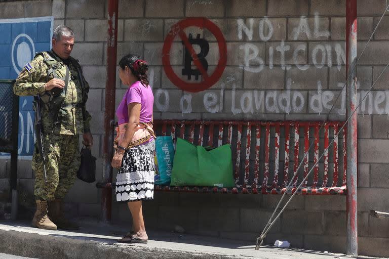 Un soldado habla con una vecina en una parada de ómnibus cubierta con graffiti en contra del uso del Bitcoin, antes de unas elecciones municipales en San José Las Flores, El Salvador