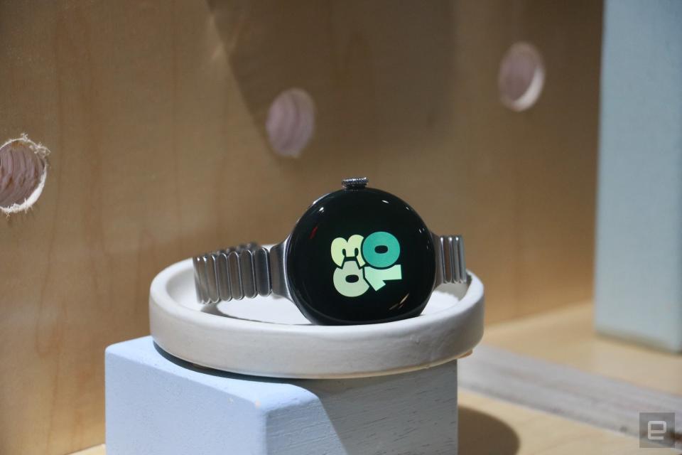 A Pixel Watch 2 with the new slim metal strap, sitting on a stand on a wooden surface. Its screen shows the time as 
