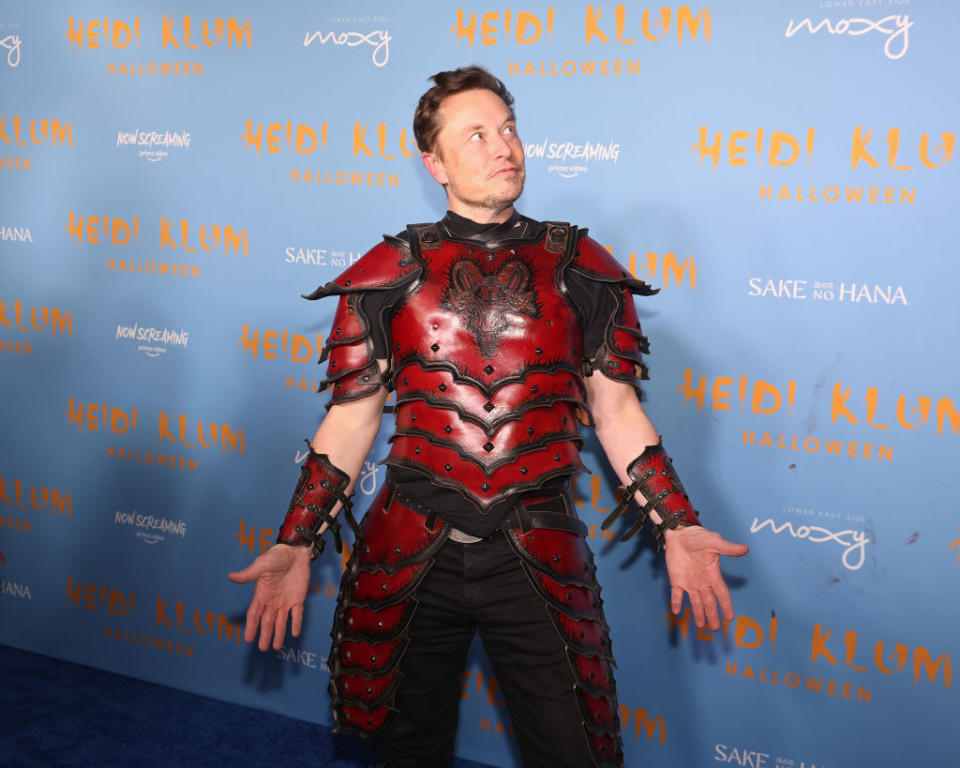 Elon Musk wearing a red medieval soldier sort of costume? There are thigh and wrist guards? He's also doing the yoga mountain pose