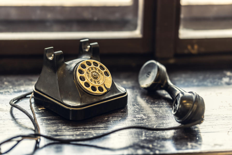 A vintage rotary telephone sits on a wooden surface with the receiver off the hook, near a window