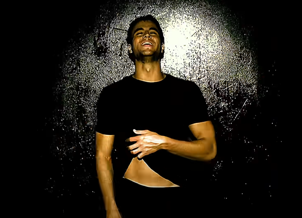 Enrique Iglesias stands against a dark background, smiling, and wearing a black t-shirt with a torn section revealing his abs