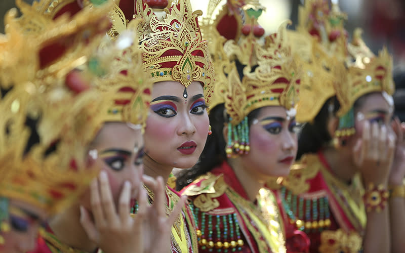 Female dancers line up before they perform during Sunday’s opening of Bali Arts Festival. They are wearing traditional outfits with gold headpieces that have red and green accents. Most of them are looking to the right, except for one woman near the center who is looking in the direction of the viewer