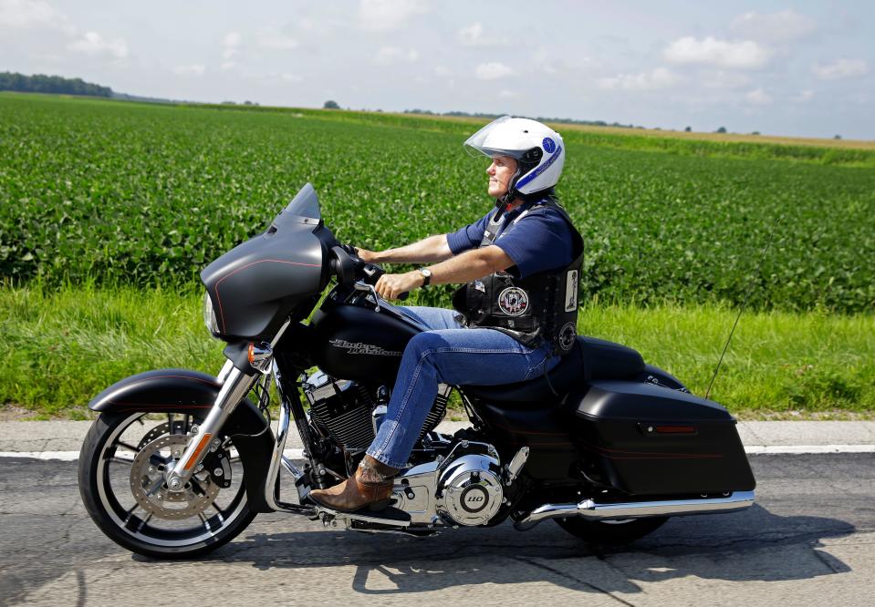 mike pence rides motorcycle past cornfields wearing a helmet, boots, jeans, and a leather vest