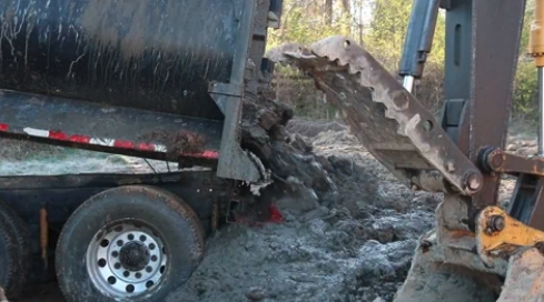 A dump truck unloads a pile of dirt, which a nearby excavator with a large bucket arm begins to spread out on the ground