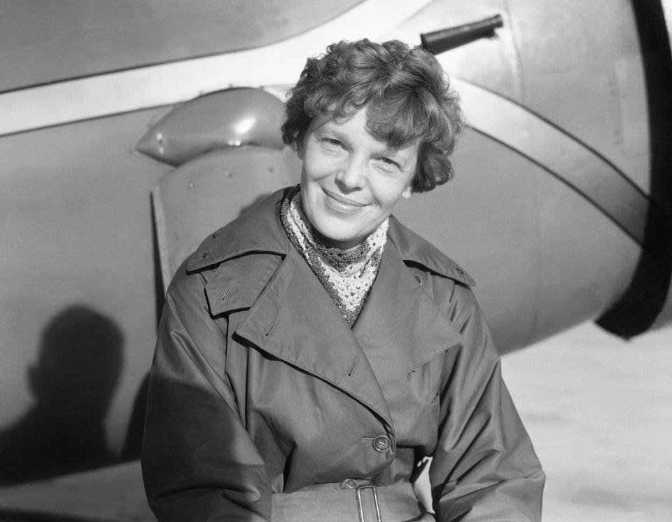 amelia earhart smiles at the camera, she is standing in front of part of a plane and wears a trenchcoat with a scarf