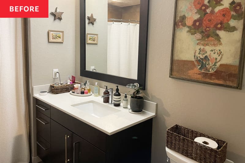 Plant and toiletries atop white sink with dark brown cabinet in bathroom with floral art on beige wall and basket for toilet paper.