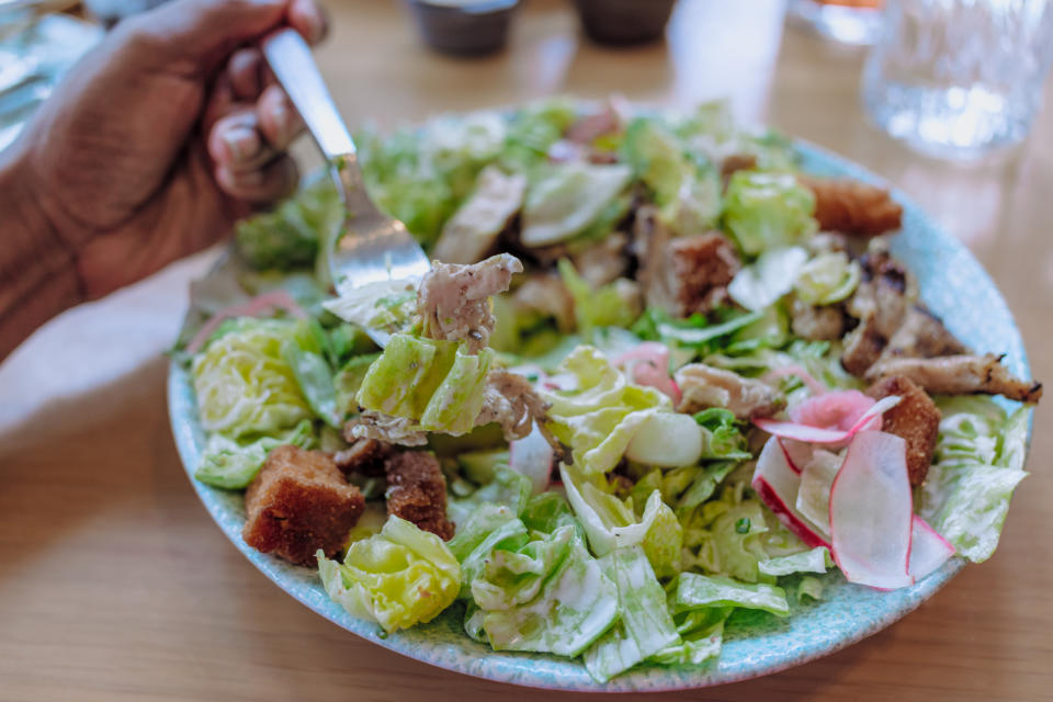 A fork holding a bite of Caesar salad above a plate filled with lettuce, croutons, radishes, and chicken