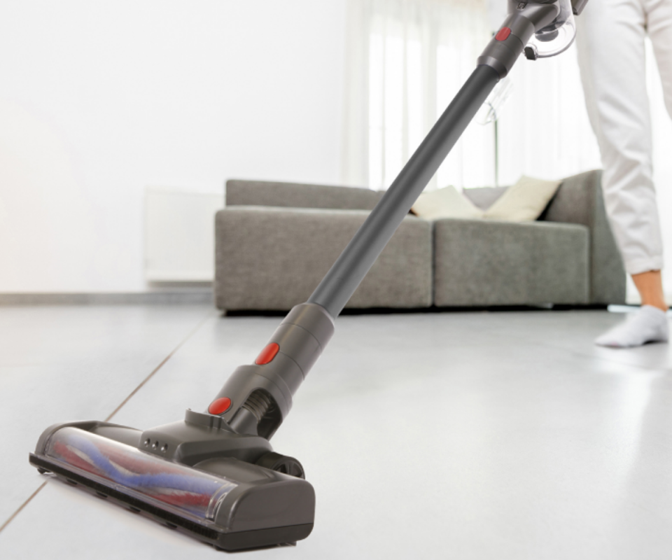 A close up of the head of the vaccum cleaning a pale floor with a woman holding it in the background with a grey lounge and a white wall.