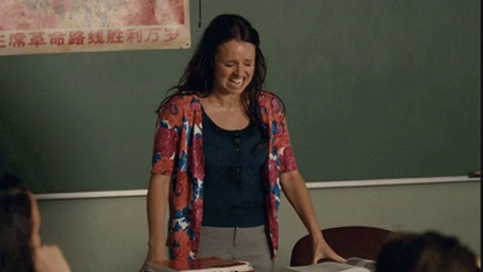 Woman in a patterned cardigan stands at a classroom desk, appearing distressed and sweating profusely