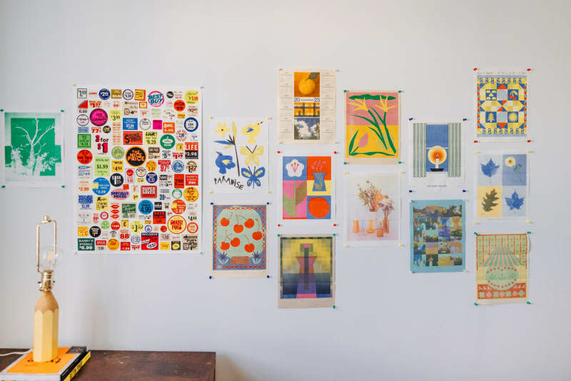 A gallery wall with colorful artwork is seen in white room.