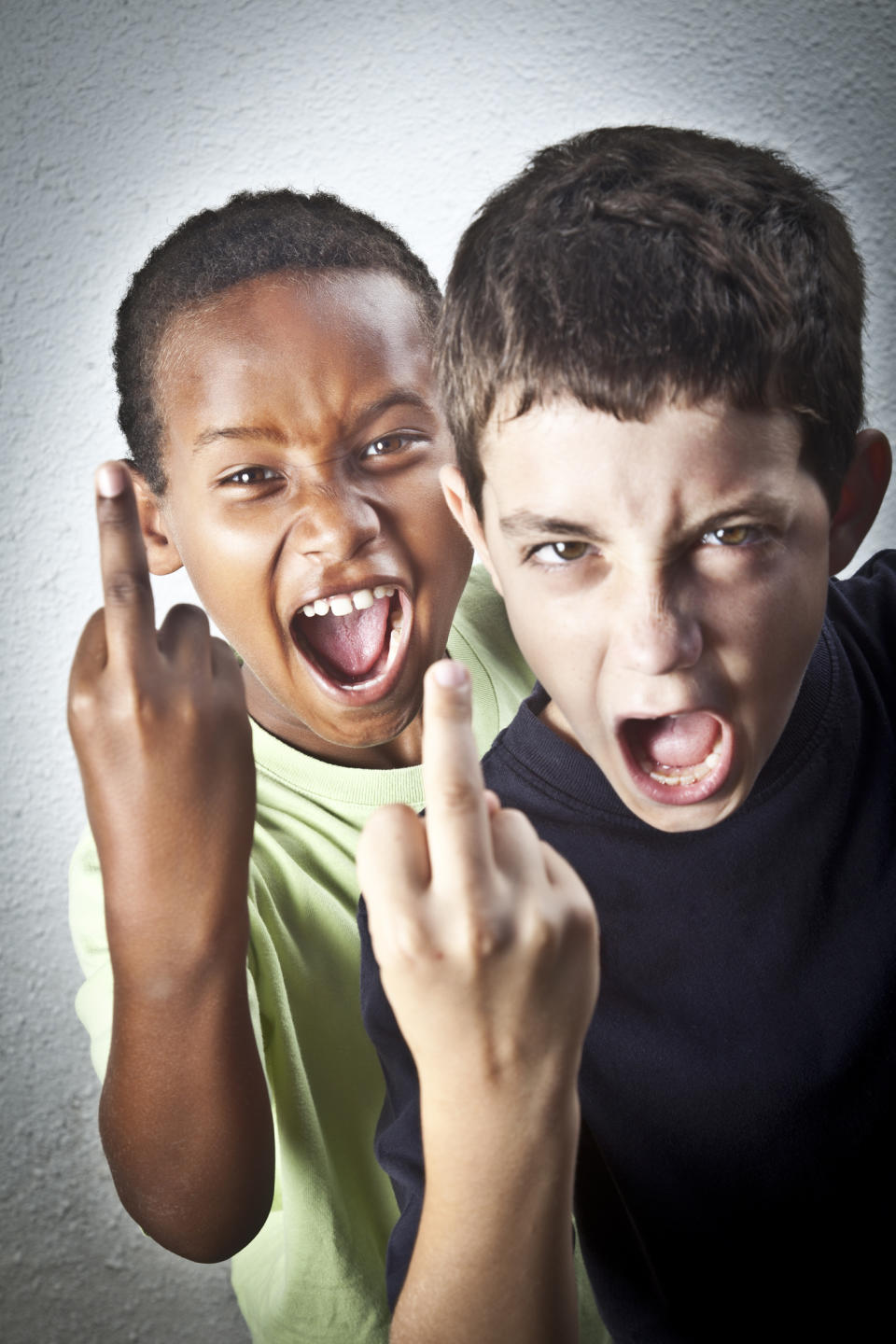 Two boys, one wearing a light shirt and the other a dark shirt, are making faces and showing their middle fingers to the camera in a playful manner