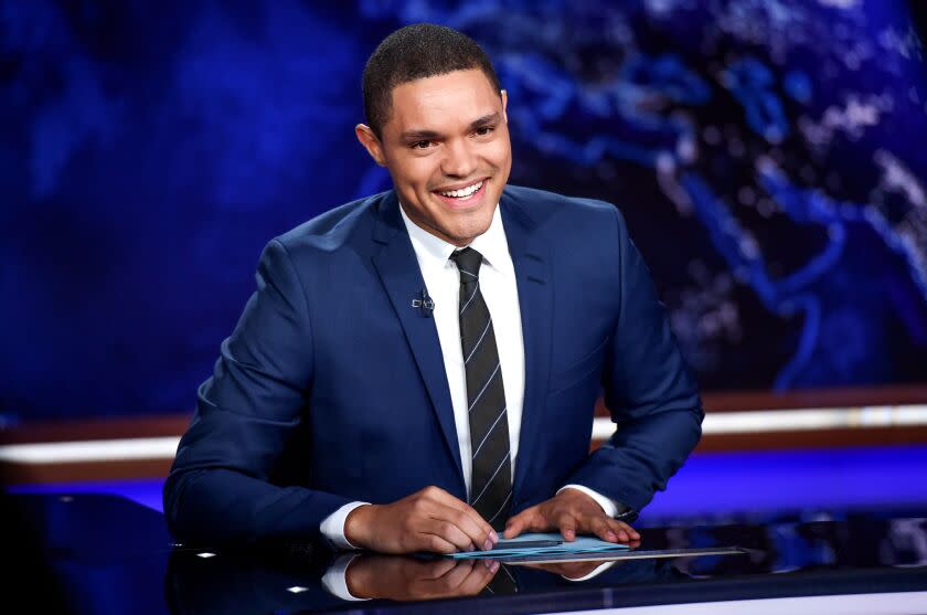 A man in a blue suit and tie smiling and sitting at a desk against a blue background