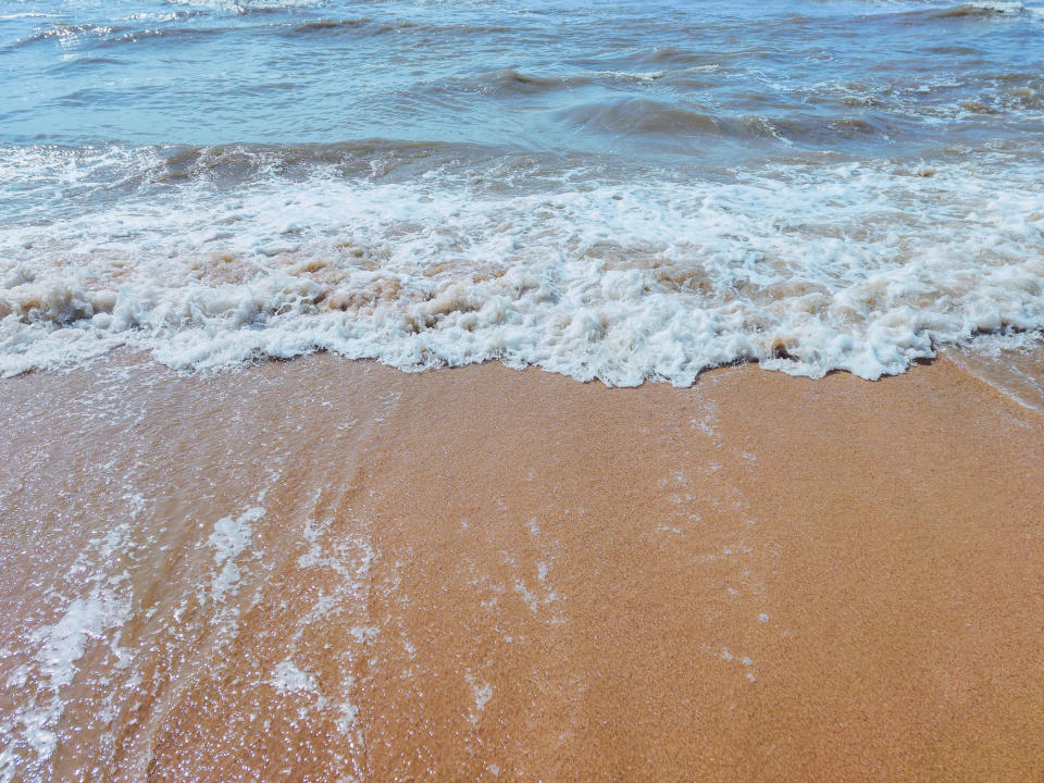 Waves gently rolling onto a sandy beach with foamy water at the shoreline. The sky is clear, and the sea is calm