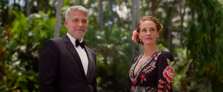 A man in a tuxedo stands beside a woman in a long gown, both looking off at something offscreen